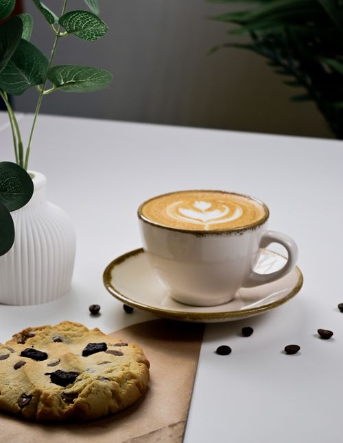 Free A cozy setup featuring a cappuccino with heart froth art, a chocolate chip cookie, and coffee beans. Stock Photo