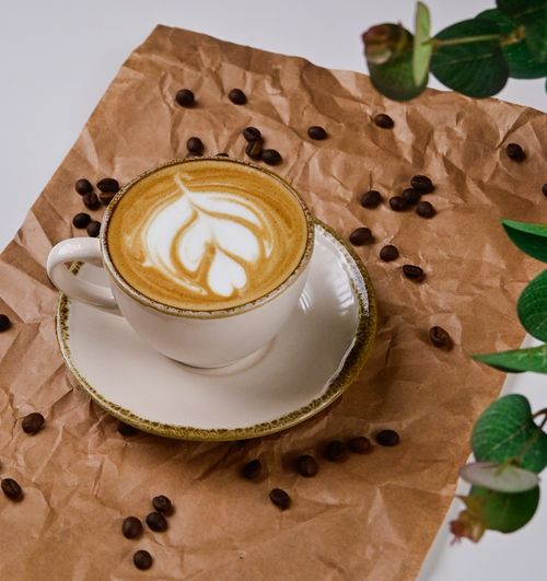 Free A beautifully crafted cappuccino with latte art sits on crumpled brown paper surrounded by coffee beans. Stock Photo