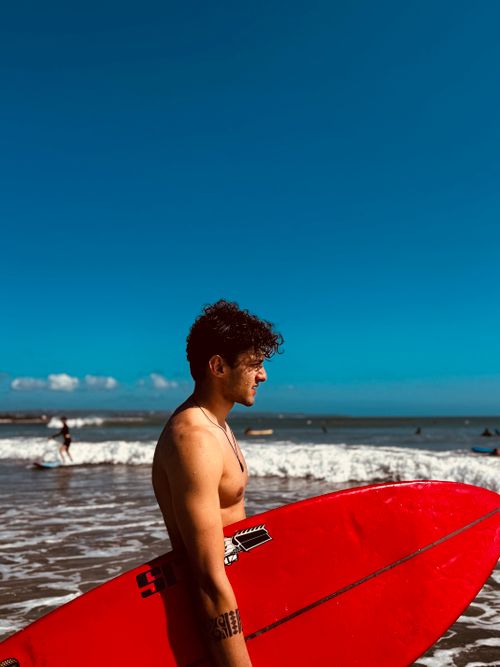 Free Side profile of a man holding a red surfboard standing at a sunny beach. Stock Photo