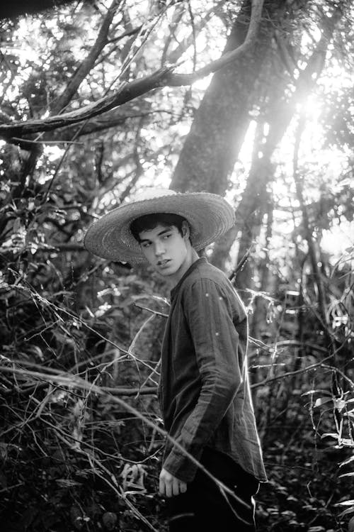Grayscale Photo of Man in Straw Hat Standing in the Middle of the Woods