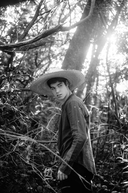 Black and white portrait of a man with straw hat standing in a sunlit forest.