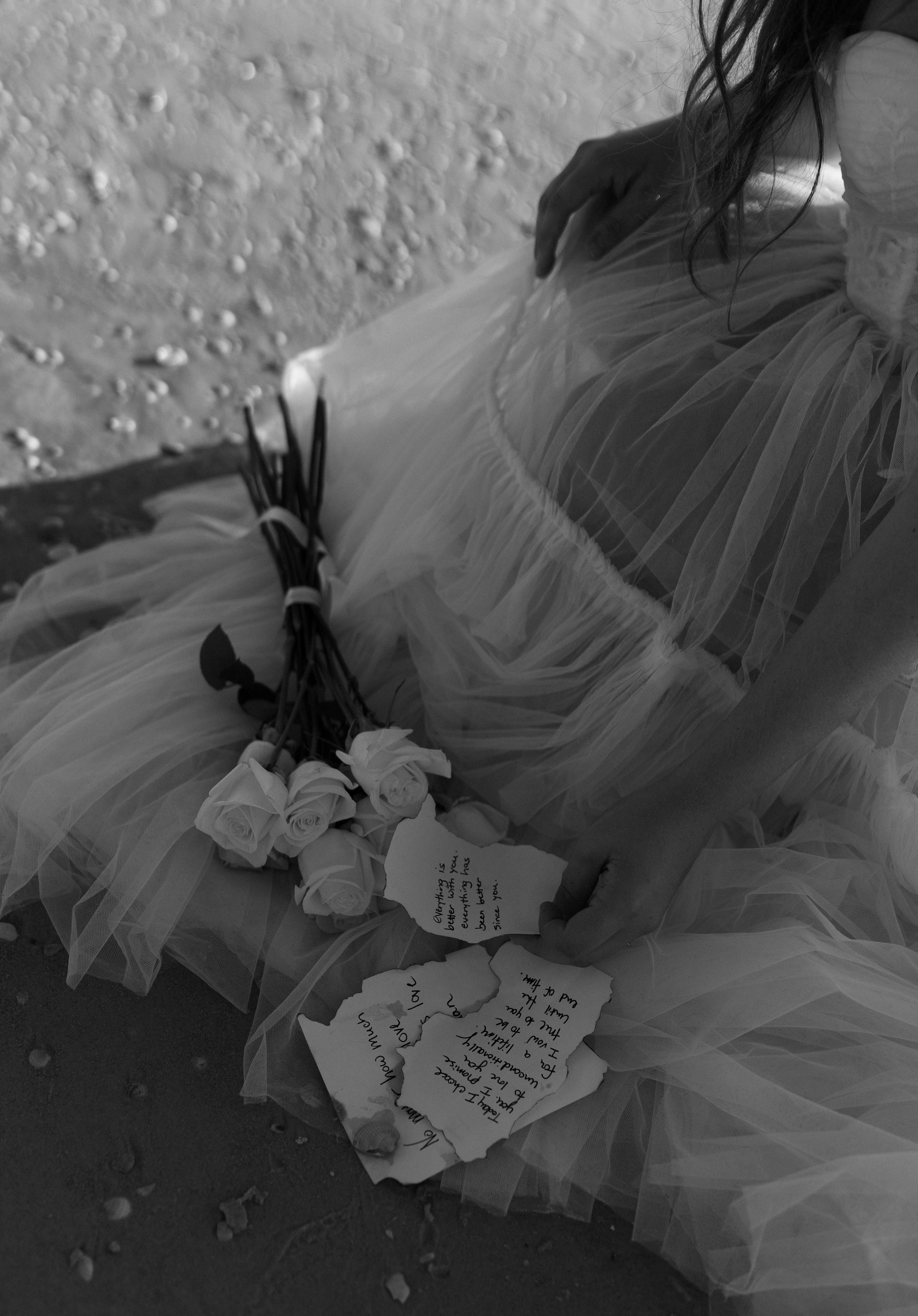 Black and white photo of a bride on a beach holding a bouquet and torn letter.