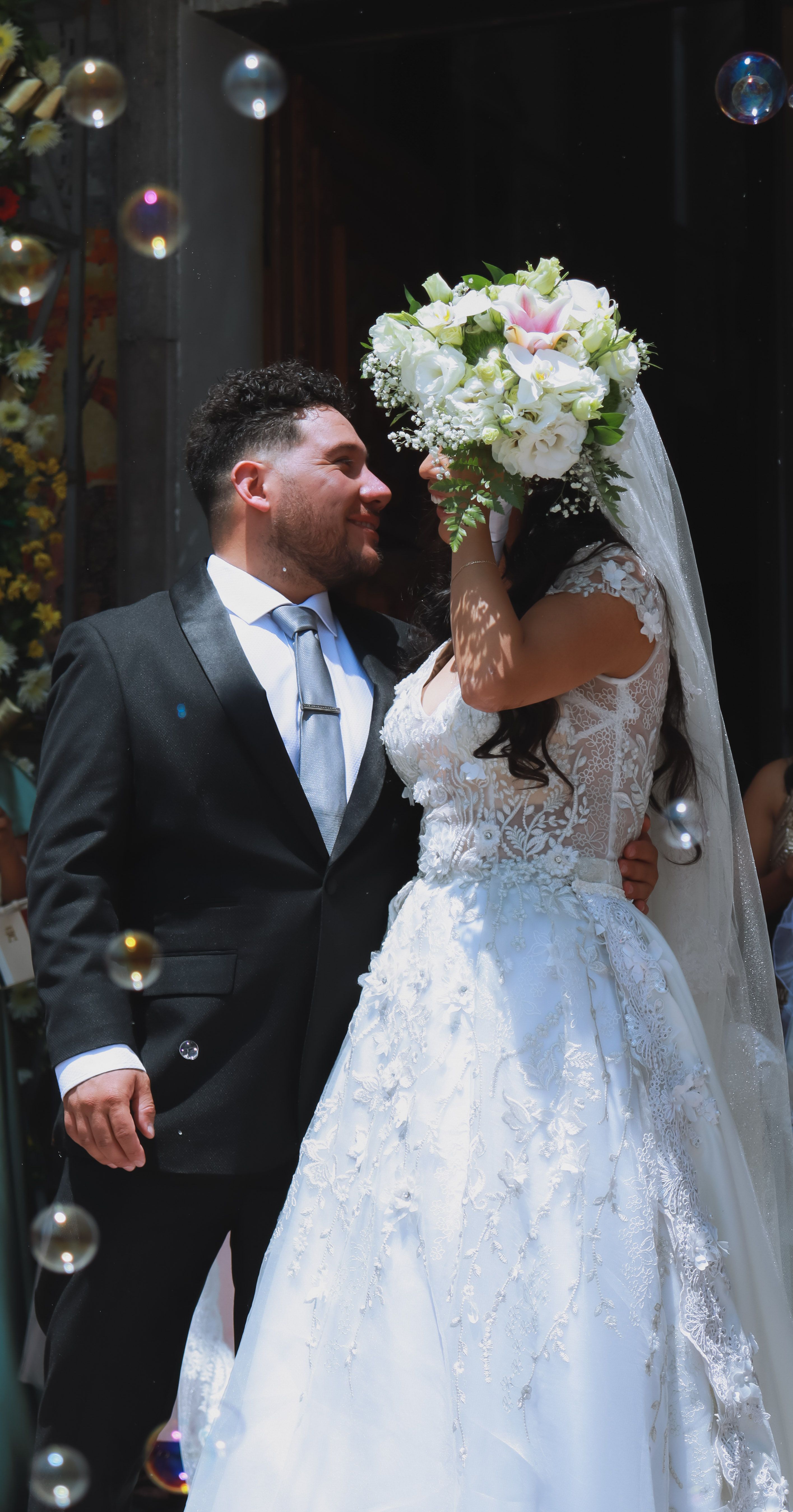 Newlywed couple embracing, surrounded by bubbles, the bride holding a bouquet tightly.