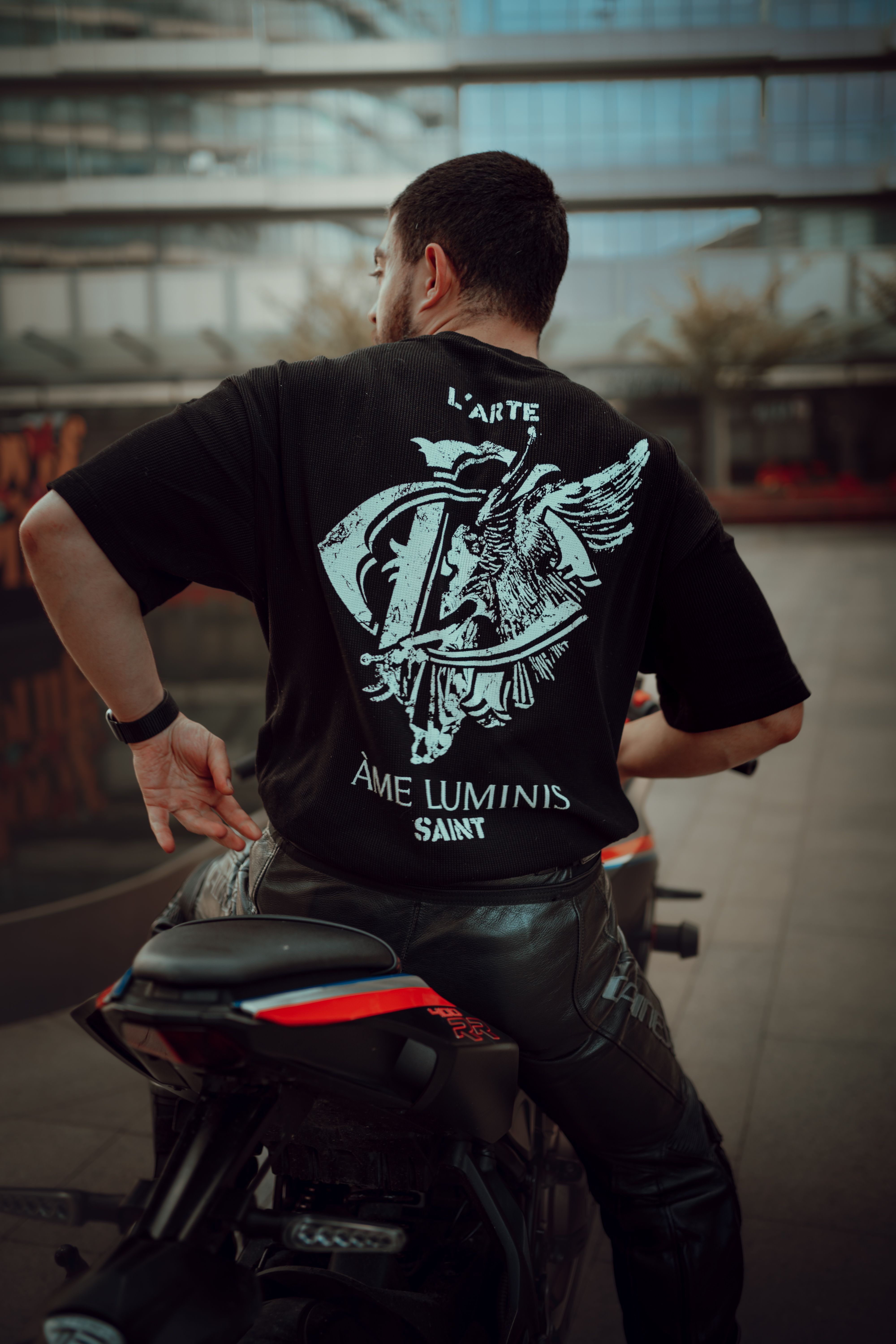 A man in black t-shirt and leather pants sits on a motorcycle in urban İstanbul.