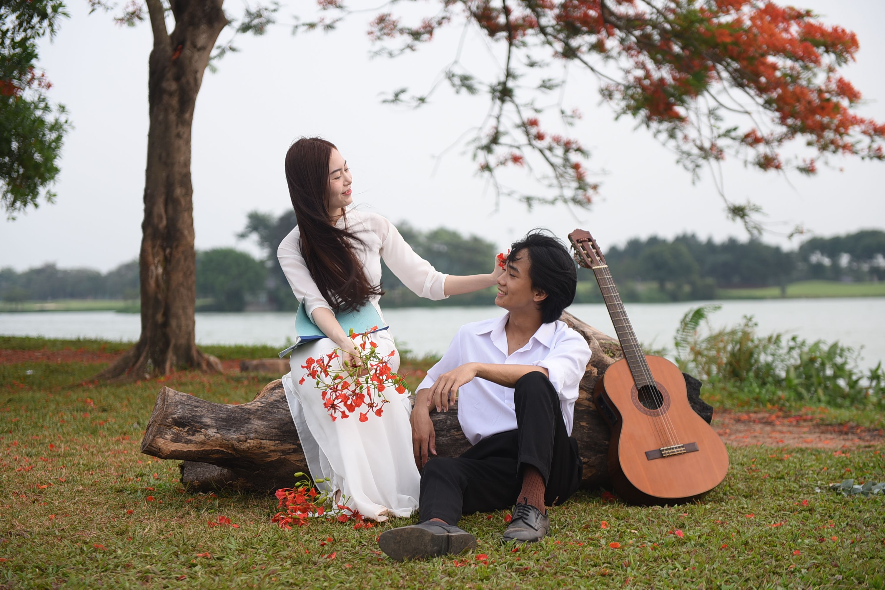 Romantic couple sitting by a tree with a guitar and vibrant flowers in a serene park setting.