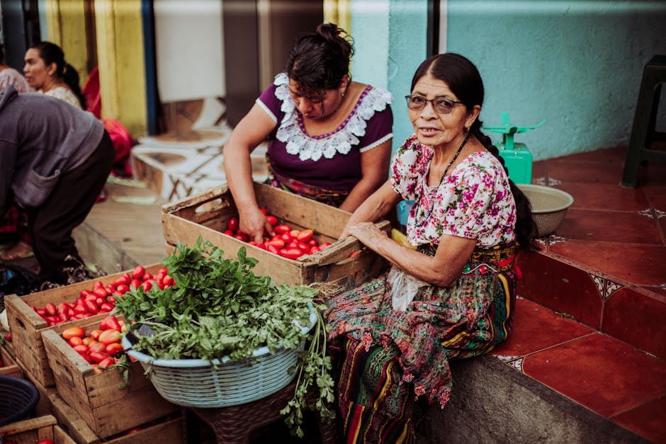 Two women in traditional attire sorting tomatoes at an outdoor market, surrounded by vibrant colors.