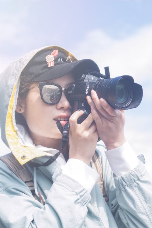 Foto De Mujer Con Chaqueta Con Capucha, Gorra Y Gafas De Sol Tomando Una Fotografía Con Una Cámara Réflex Digital Nikon Negra