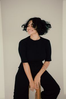 A young woman with tattoos smiling while sitting on a stool indoors.