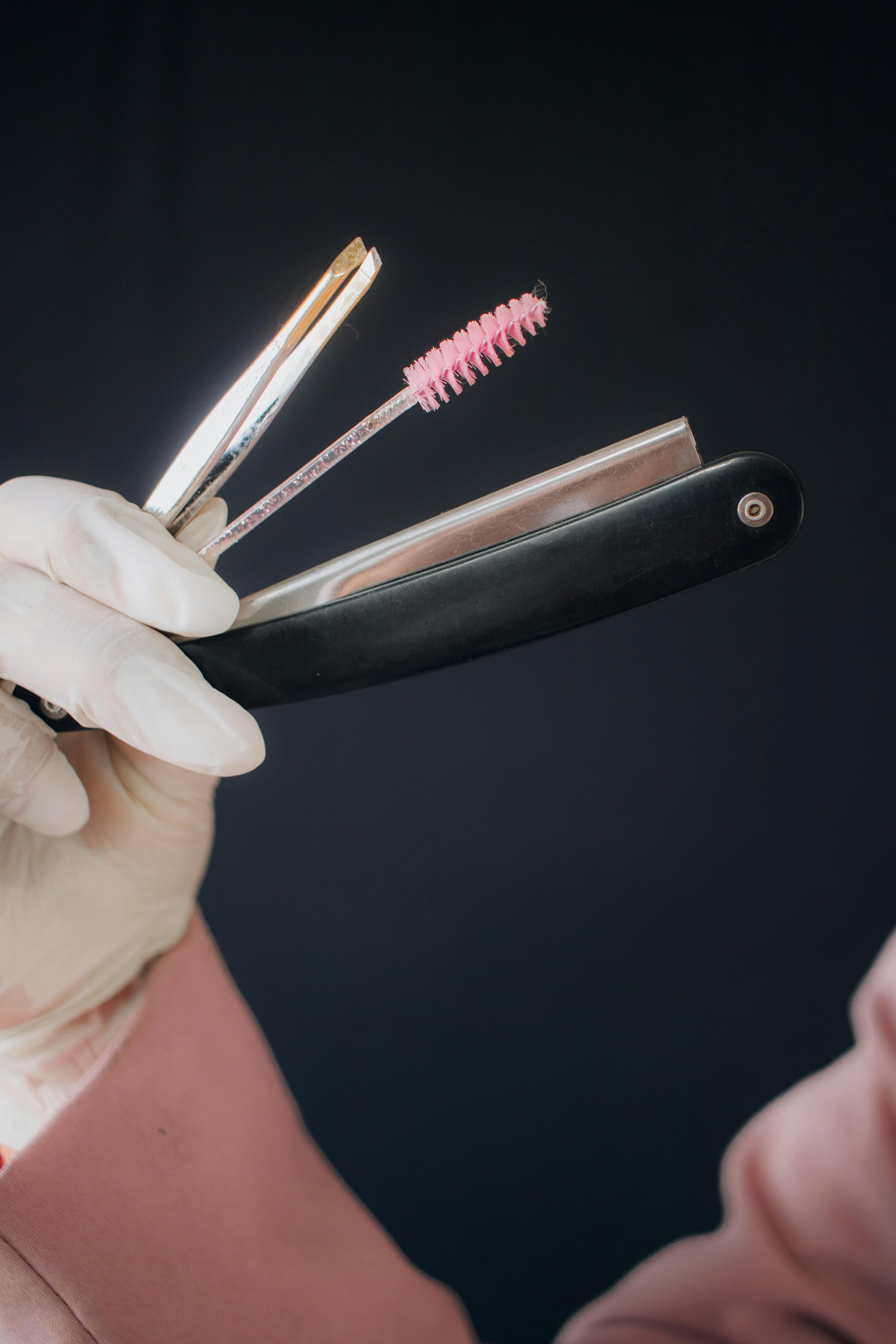 Gloved hand holding straight razor and pink brush against dark background, emphasizing hygiene and precision.