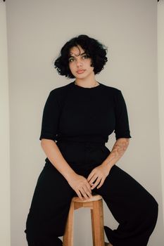 Portrait of a young woman with tattoos sitting on a stool against a white background.