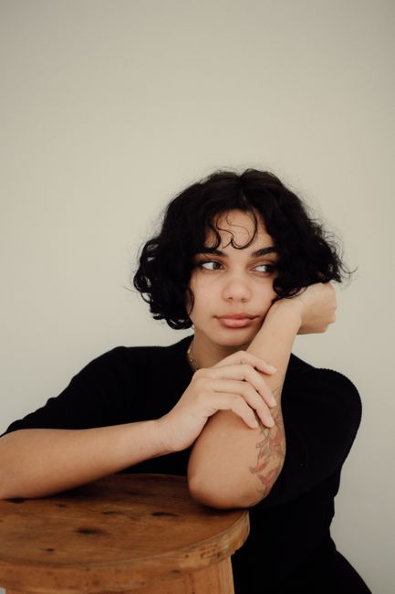 Young woman in a black t-shirt leaning on a stool, looking pensive against a white background.