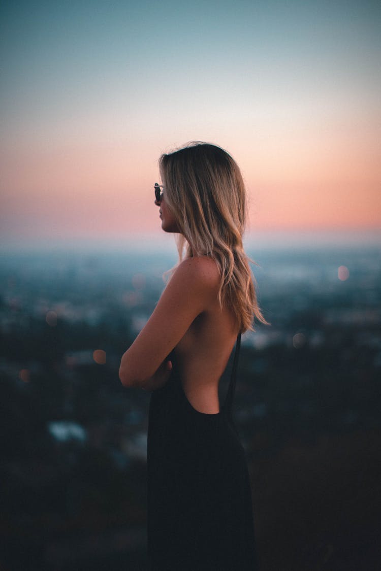 Side View Photo Of Woman In Black Dress Standing With Her Arms Crossed Looking Into The Distance.