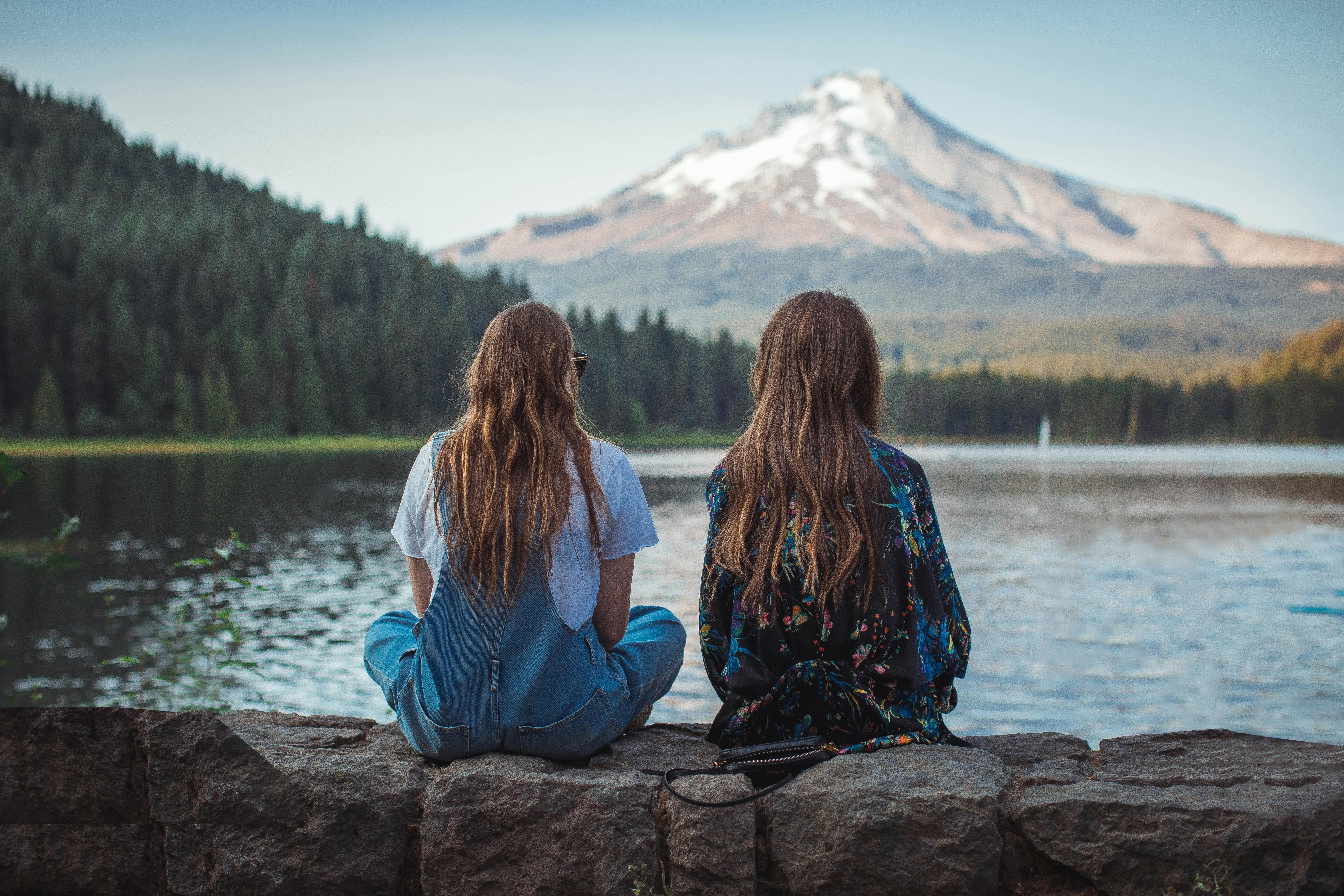 two women sitting on rock facing on body of water and mountain