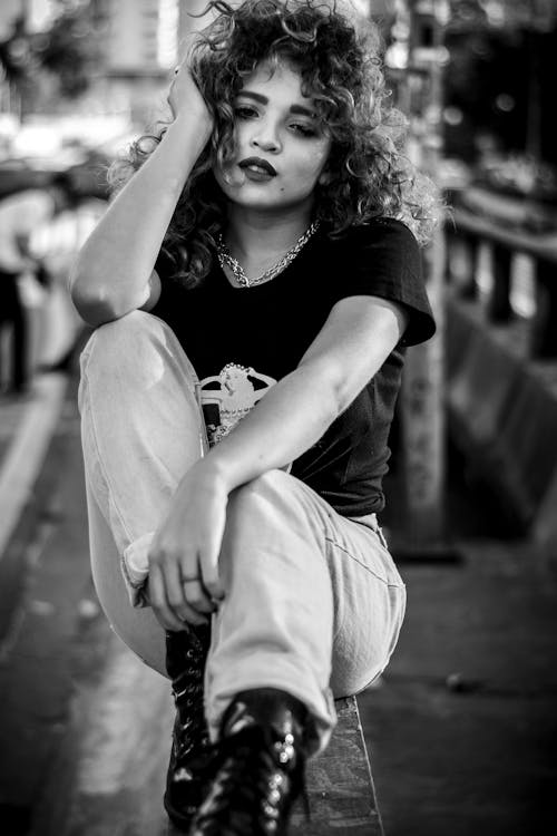 Grayscale Photo of Woman in T-shirt, Pants, and Boots Sitting on Railing Posing