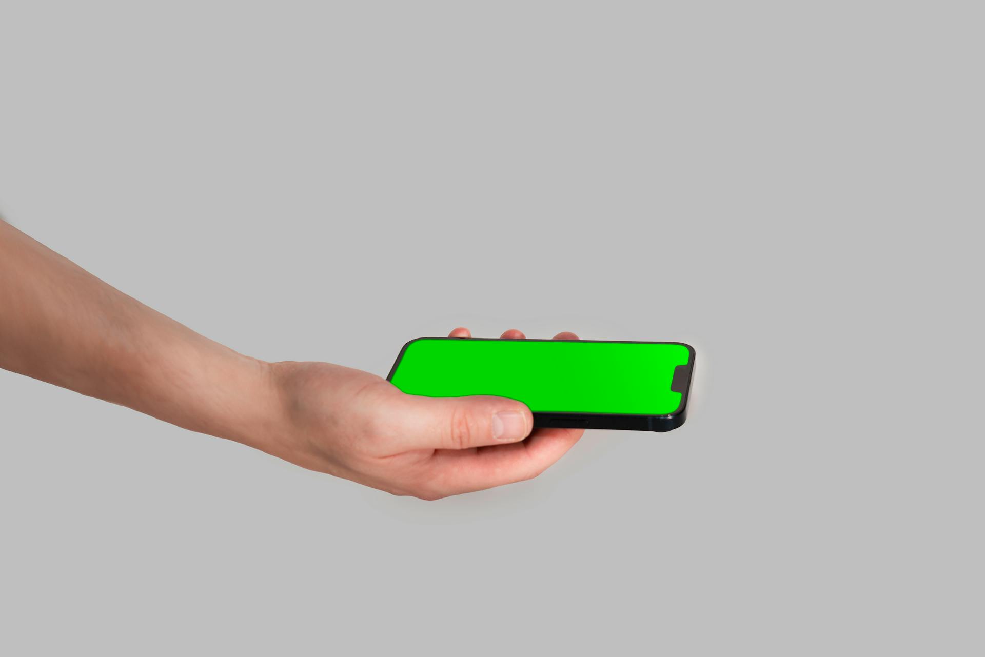 A hand holding a smartphone with a vivid green screen on a gray background, perfect for mockup use.