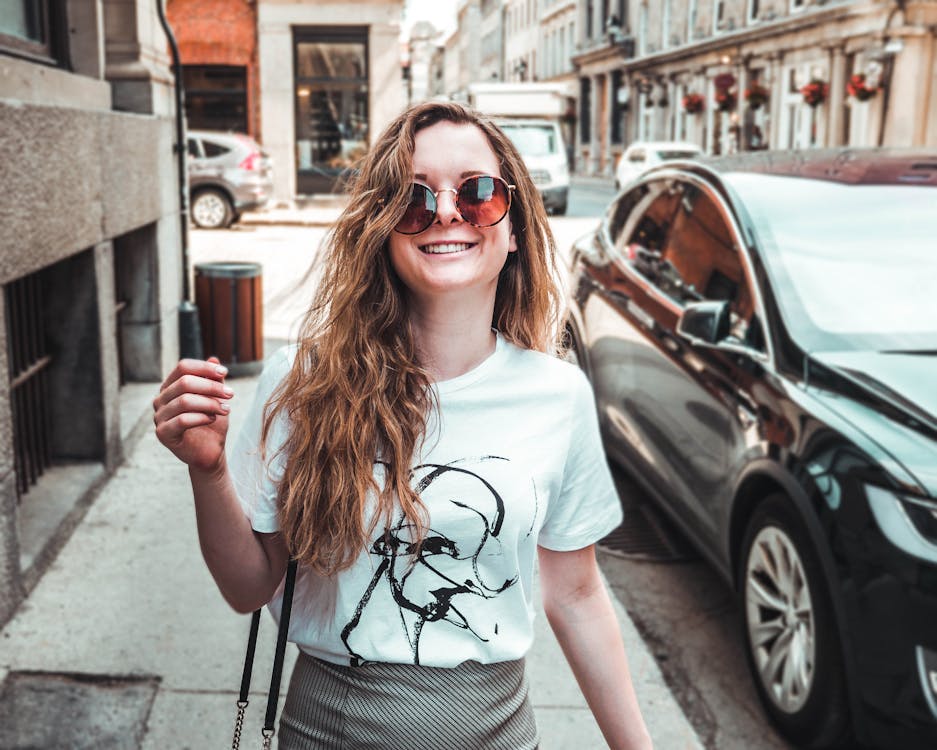 Photo of Smiling Woman in White T-shirt and Sunglasses Walking on Side Walk