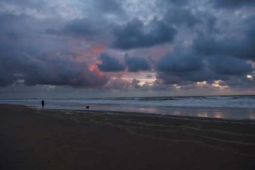Silhouetted person walking dog along a peaceful beach with dramatic sunset clouds overhead.