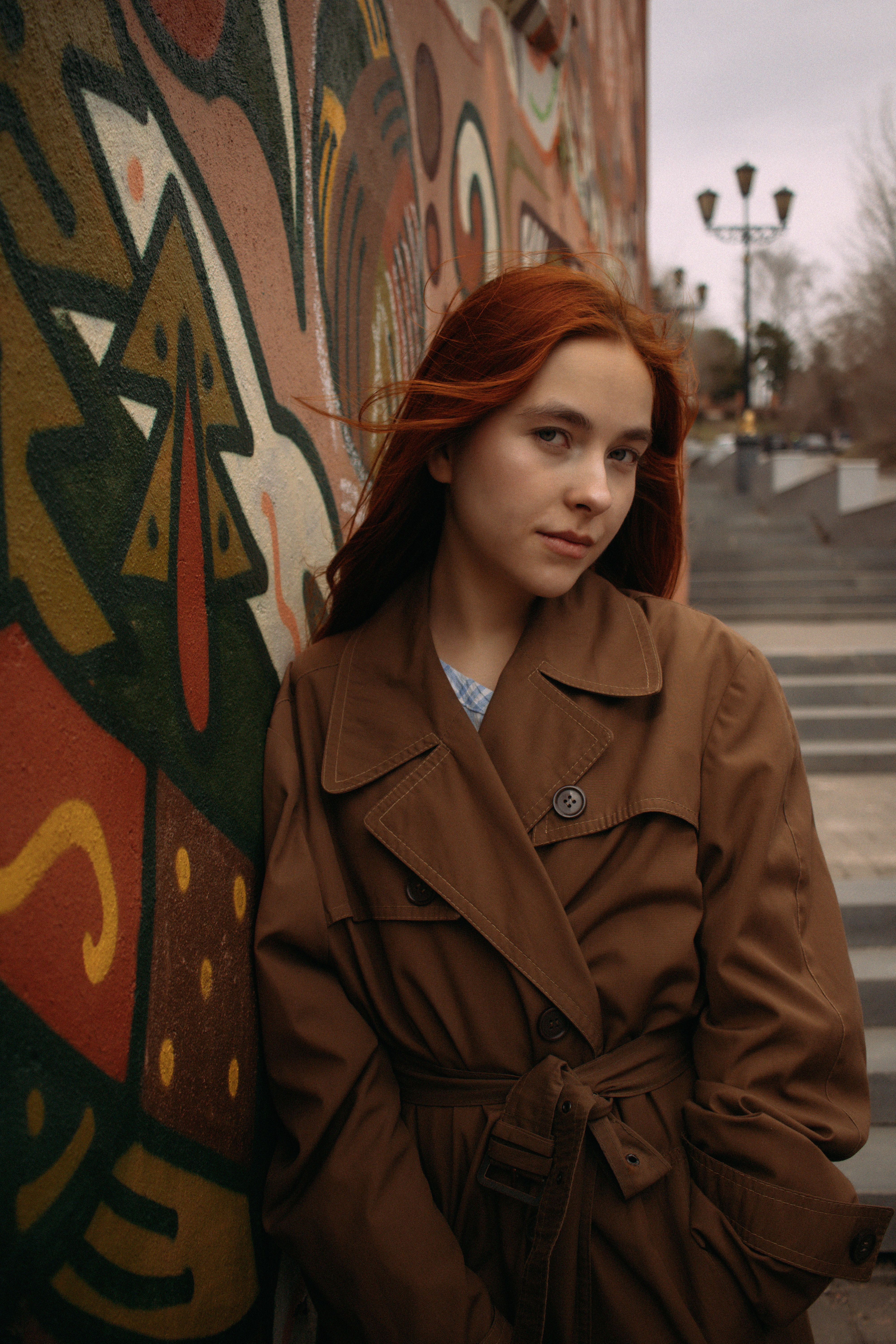 Redheaded woman in trench coat posing by a colorful graffiti wall in urban Tomsk.