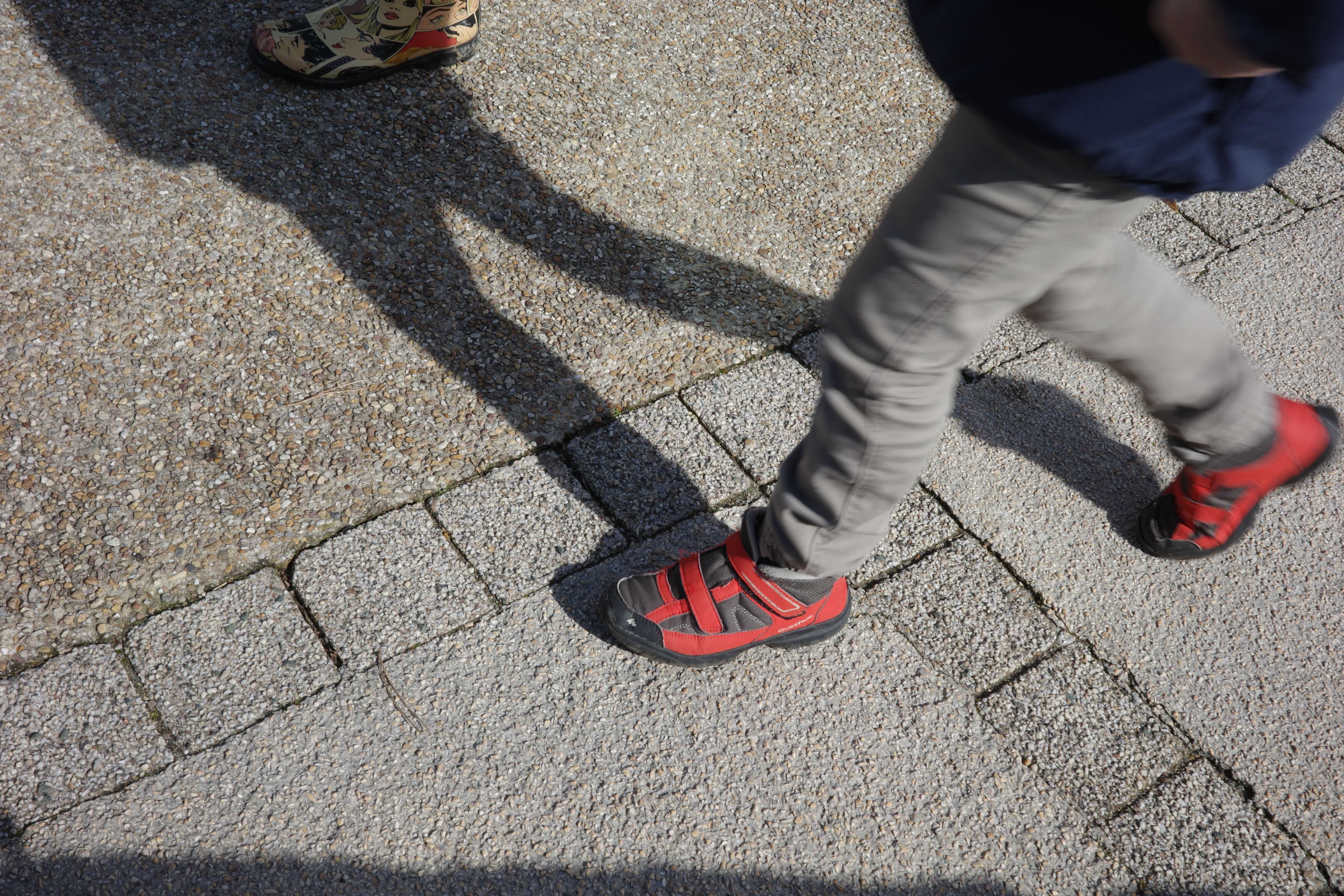 A child wearing red shoes walks on a city pavement. Shadows create a dynamic pattern.