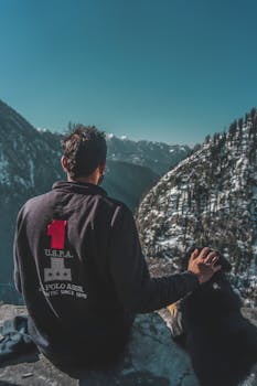 一名男子和他的狗坐在悬崖上，背影俯瞰着雪山