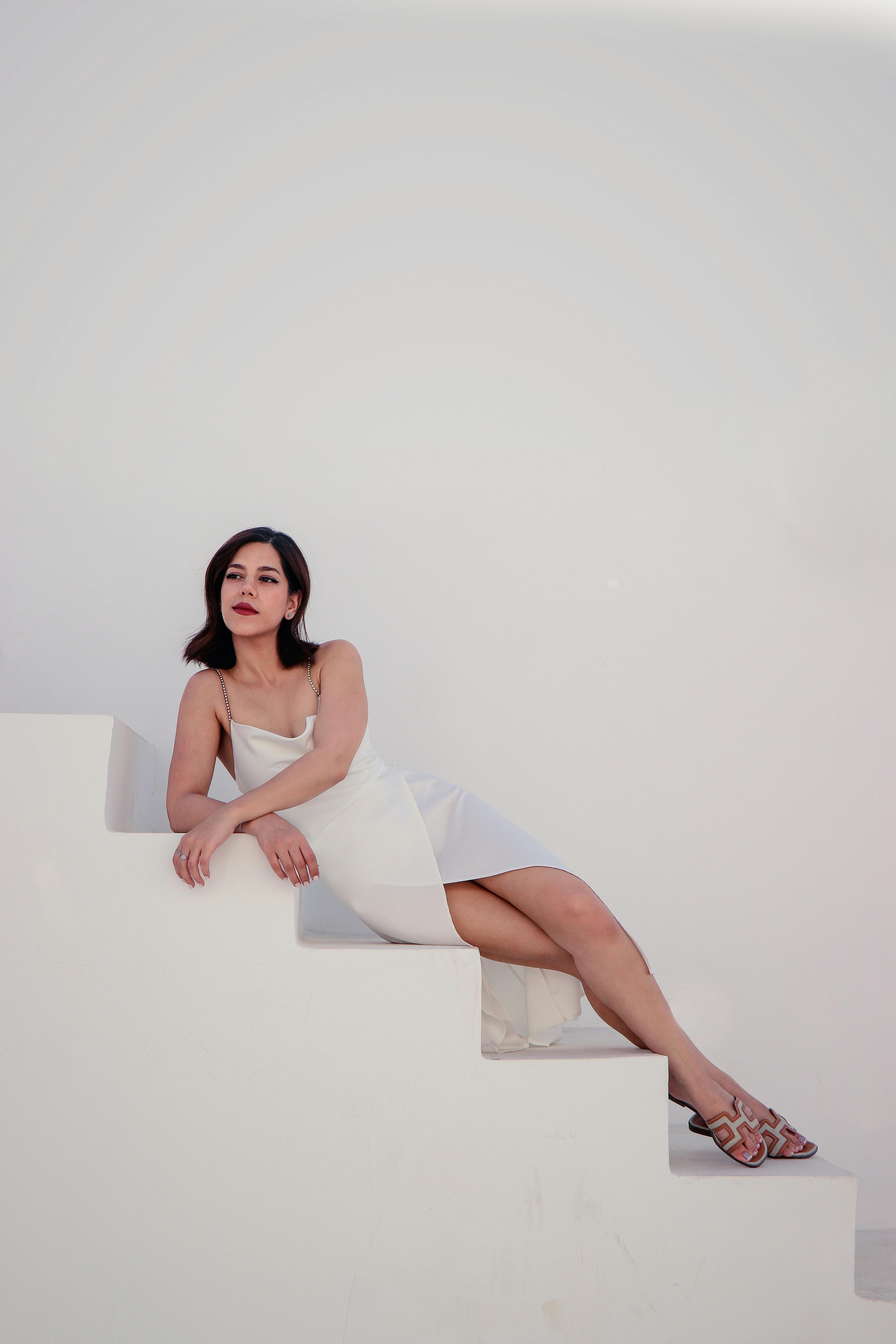 A stylish woman in a white dress elegantly sitting on minimalist stairs against a white background.