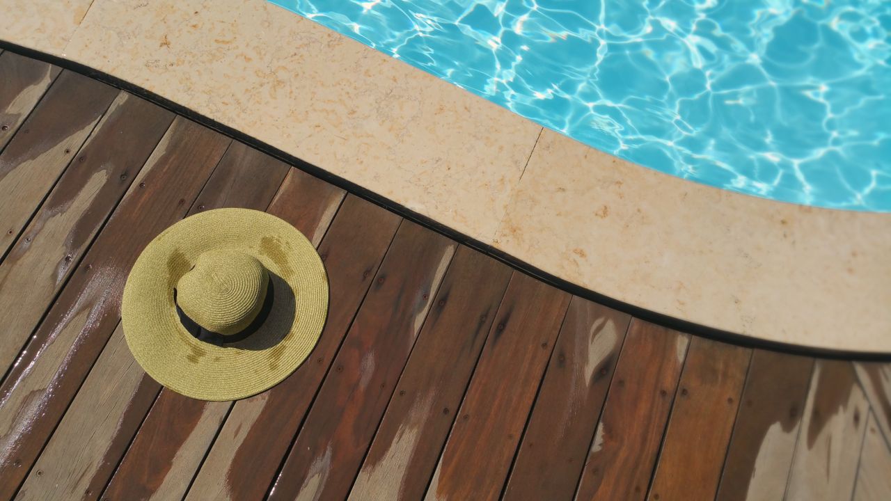 a straw hat rests on a wooden deck beside a sparkling blue swimming po