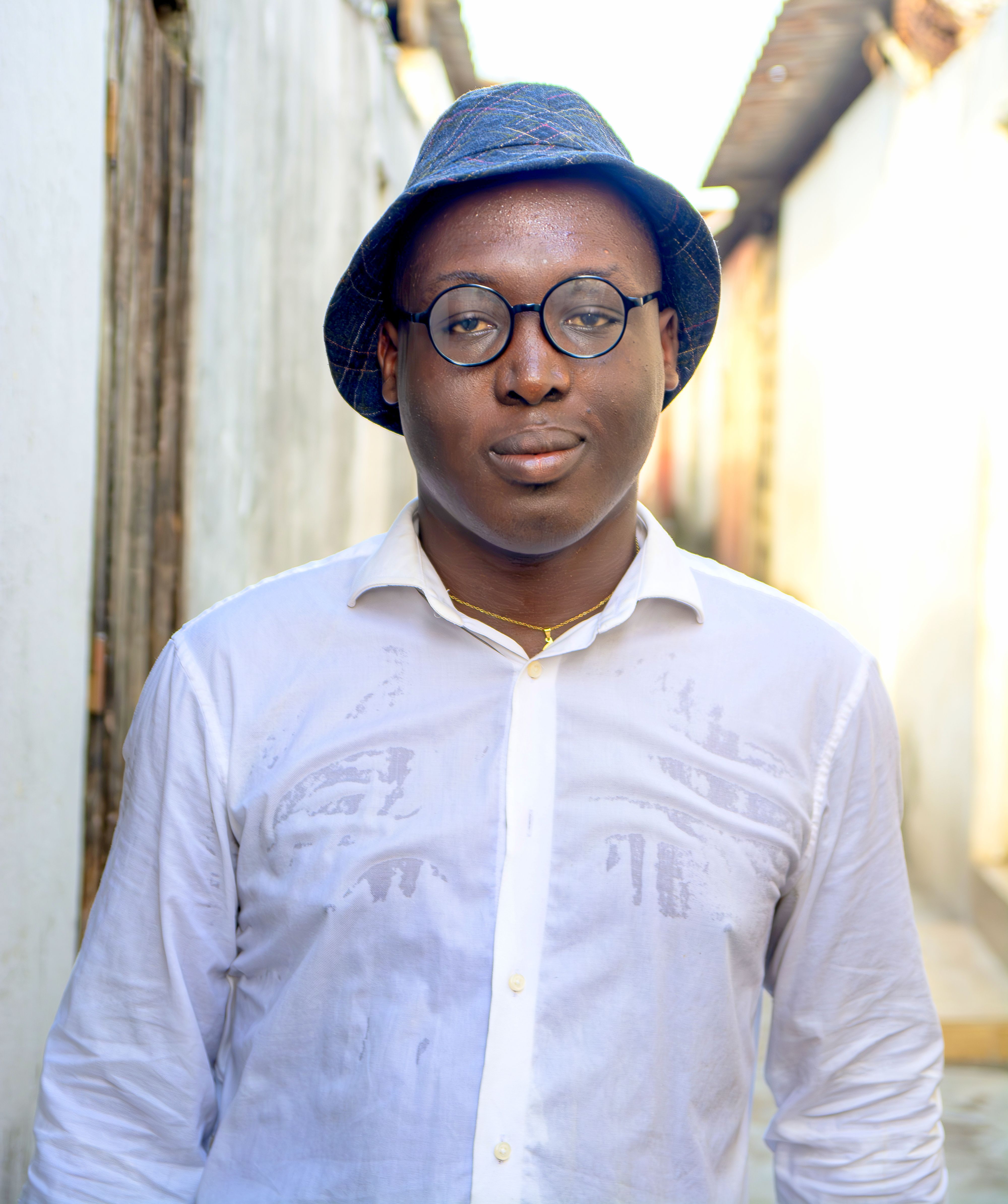 A man wearing glasses and a hat poses on a narrow city street, exuding a casual and confident vibe.