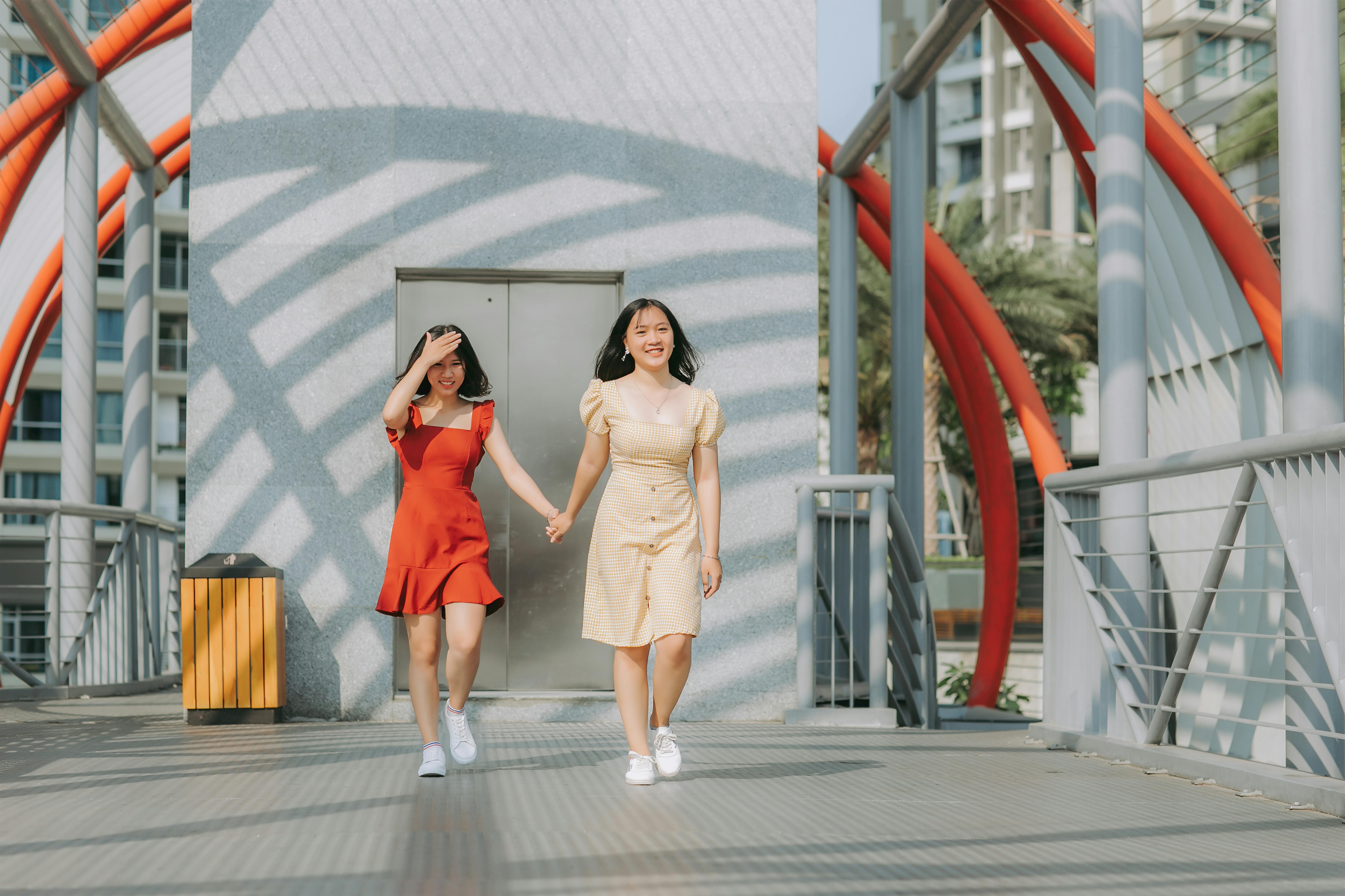 两个女人手牵着手，面带微笑，走在一座现代桥梁上，桥上有红色拱门，背景是城市