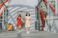 两个女人手牵着手，面带微笑，走在一座现代桥梁上，桥上有红色拱门，背景是城市