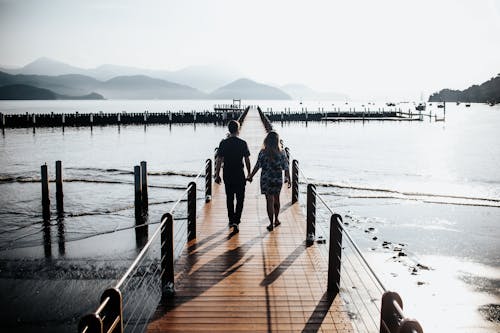 Back View Photo of Couple Holding Hands While Walking Down Wooden Dock