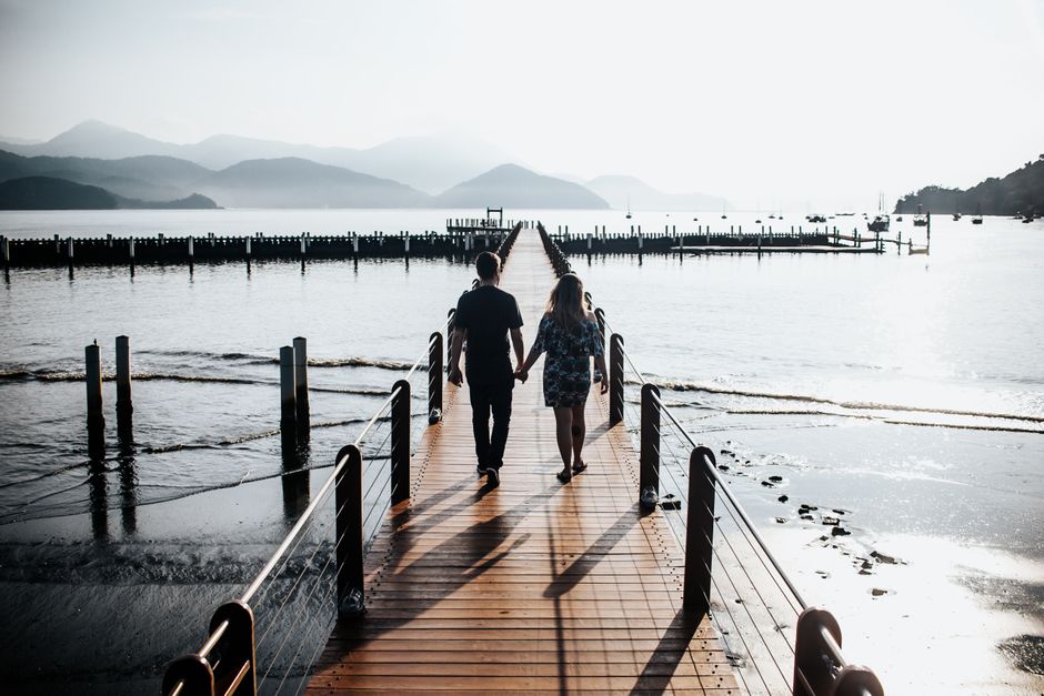A couple holding hands walking along a pier overlooking a serene lake.