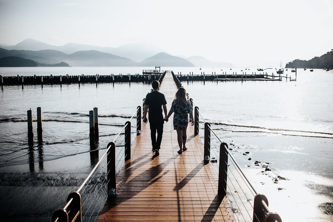 Back View Photo of Couple Holding Hands While Walking Down Wooden Dock