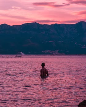 A lone figure in Montenegro's scenic lake at sunset, silhouetted against vibrant twilight hues. by Rakicevic Nenad