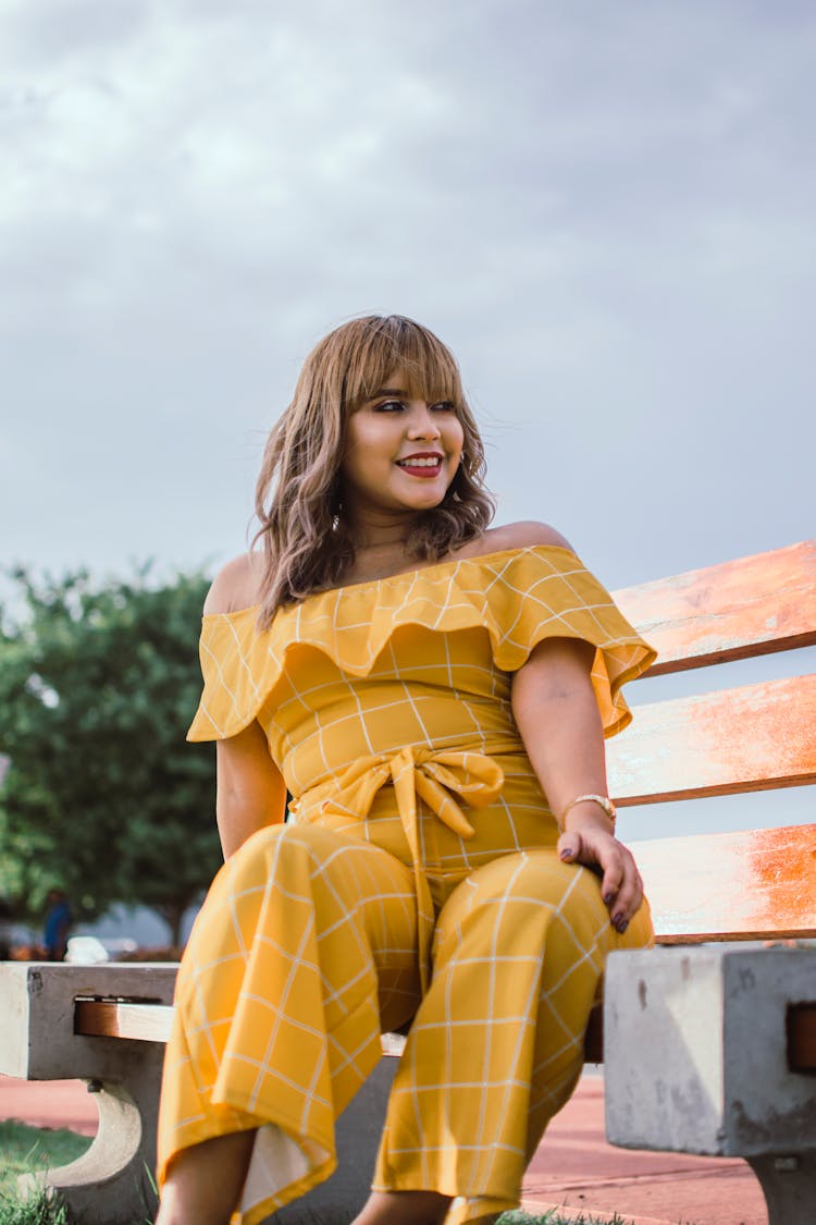 Photo Of Smiling Woman In Yellow Jumpsuit Sitting On Wooden Bench Posing While Looking Away