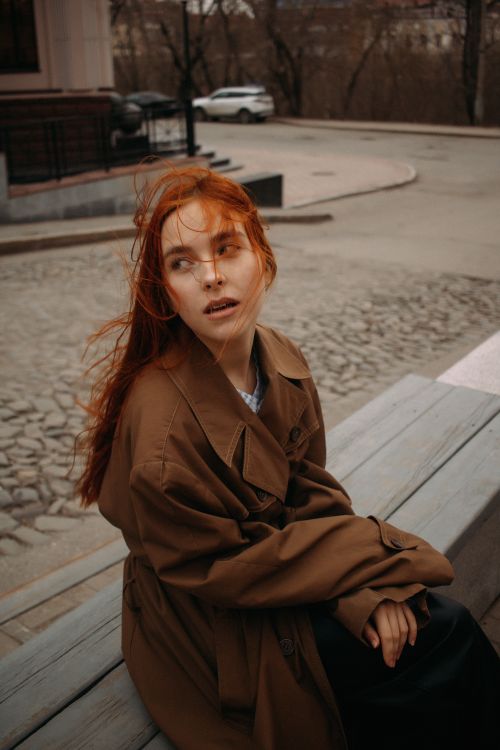 Free A woman with red hair wearing a trench coat sits on an outdoor bench in an urban area during autumn. Stock Photo
