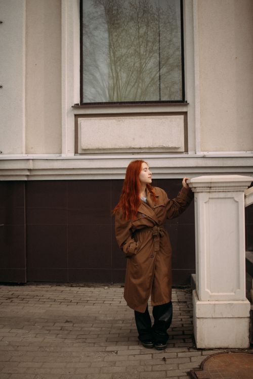 Free A redhead woman in a brown coat poses against a small column in an urban environment. Stock Photo