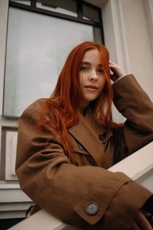 Free Stylish young woman with red hair poses confidently in a brown coat against a city backdrop. Stock Photo