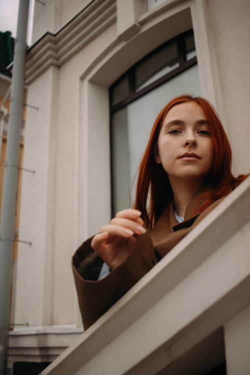 Free A thoughtful young redhead woman stands by a window, exuding a calm presence in an urban setting. Stock Photo