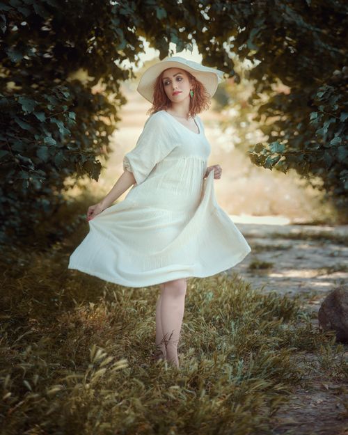 Free Woman in white dress with hat posing gracefully outdoors surrounded by lush greenery. Stock Photo