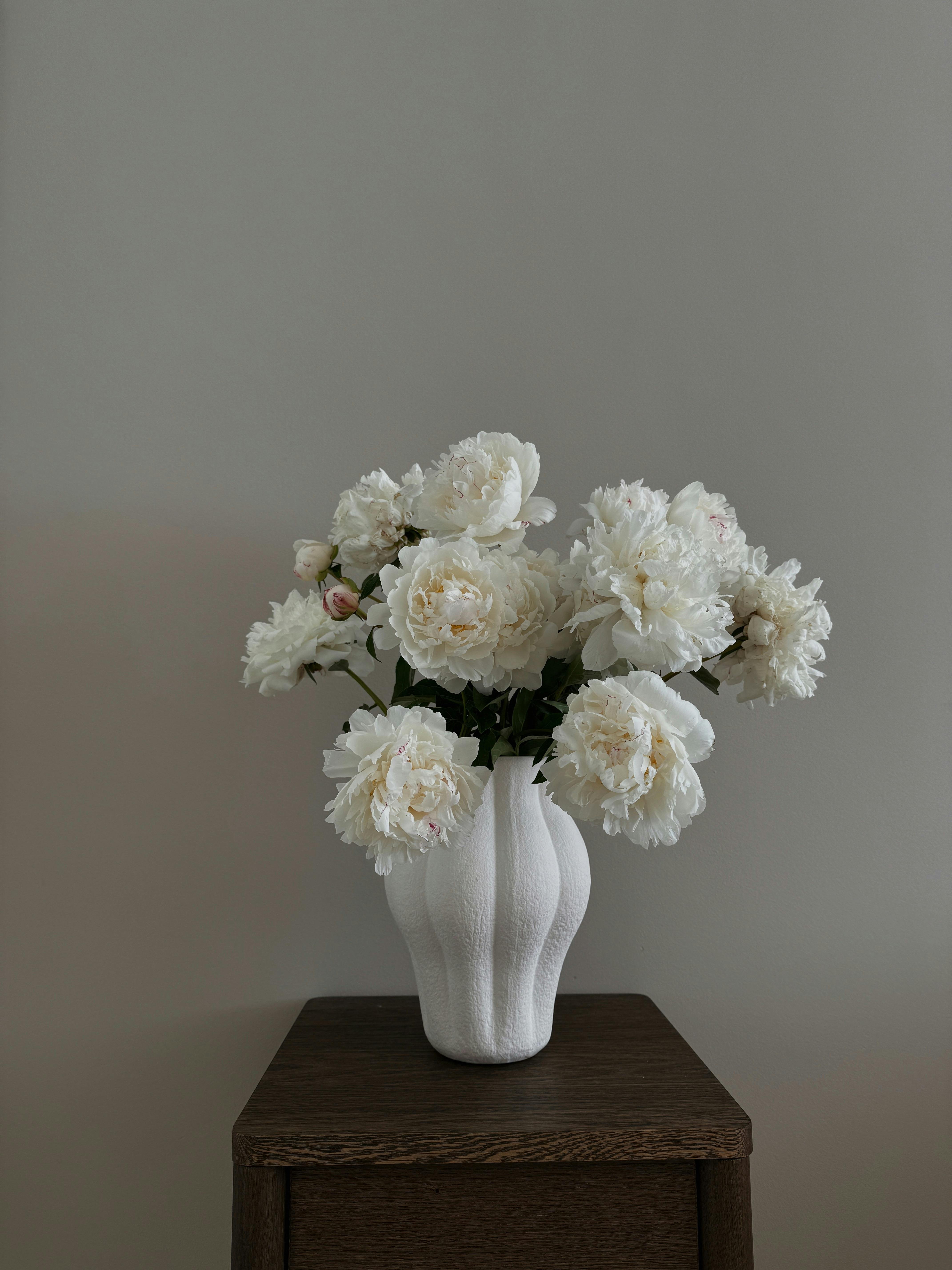 A stunning arrangement of white peonies in a textured vase on a dark wooden surface, perfect for elegant decor.