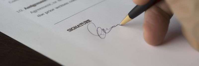 A hand signs a formal contract with a pen on a wooden desk.