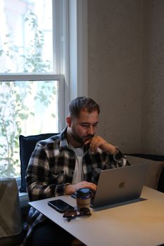 Man in plaid shirt working on laptop near window, indoors with coffee.