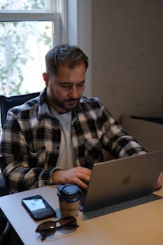 Adult man in checkered shirt using laptop and smartphone at a desk near window.