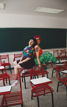 Two young women with colorful styles sitting on desks in a modern classroom.