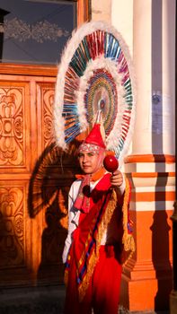 A vibrant traditional performer with maraca in colorful attire stands out in sunlight against a decorated wooden door. — photo by Anya  Juárez Tenorio