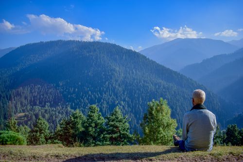 Free A senior man sitting, enjoying the panoramic view of lush forested mountains under a clear blue sky. Stock Photo