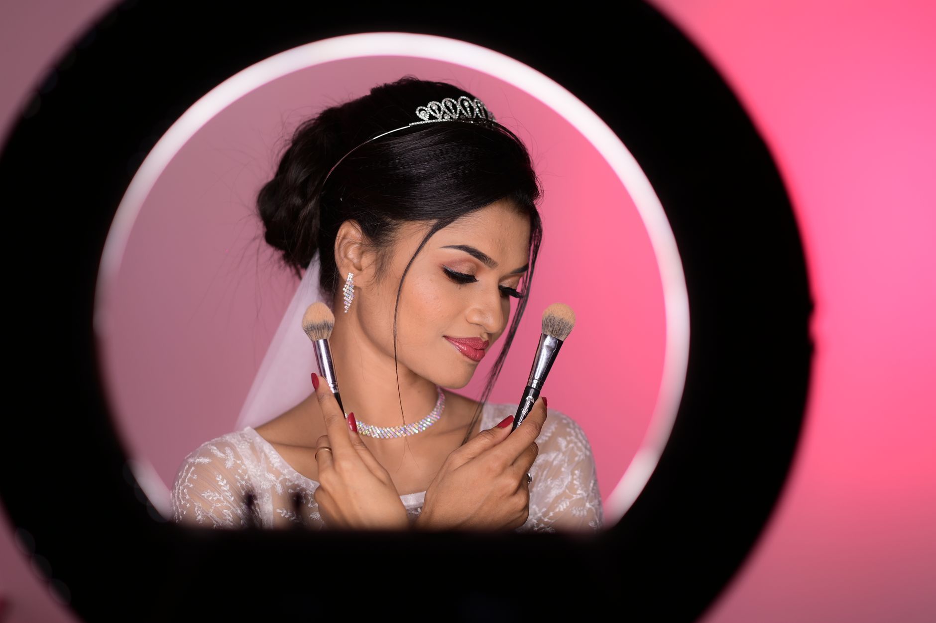 A beautiful bride with closed eyes holds makeup brushes, set against a vibrant pink background.