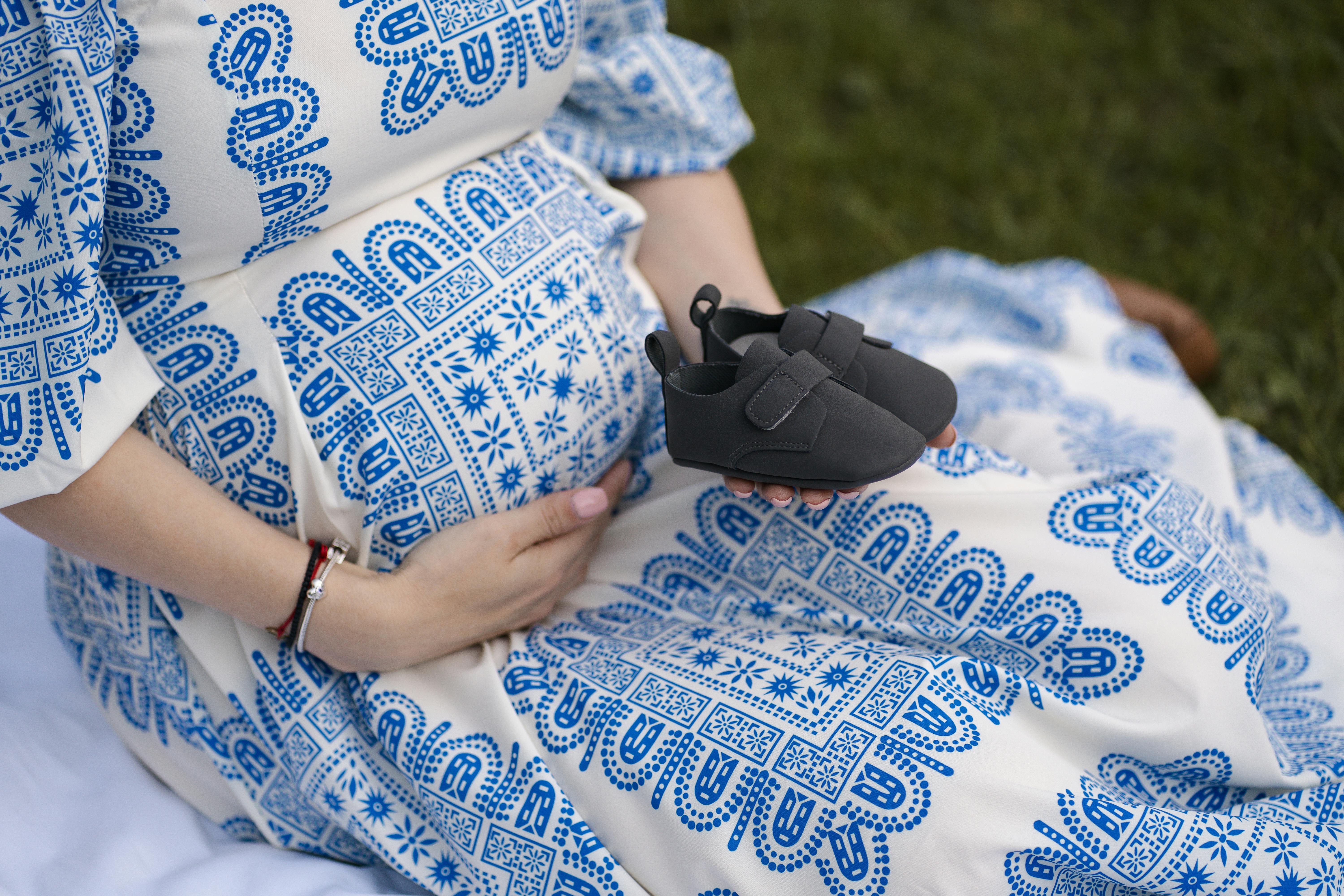 Close-up of pregnant woman in blue and white dress, holding baby shoes.