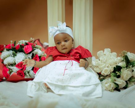 A cute baby dressed in a red and white outfit sits among beautiful bouquets, perfect for celebrations.