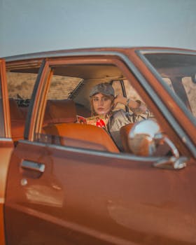 A young woman wearing a baseball cap, sitting inside a vintage car, captured in a casual and thoughtful portrait.